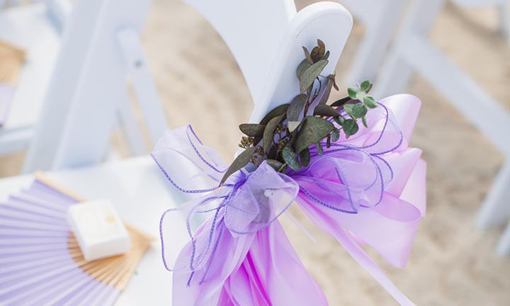 Elegant Wedding Décor Setup on a Royal Caribbean Beach Ceremony A white ceremony chair on a beach decorated with a purple ribbon and greenery for a Royal Caribbean wedding experience.